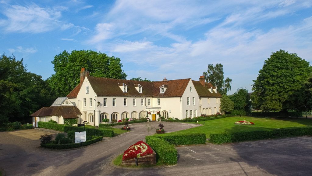 A large, historic white building with a red-tiled roof and arched windows sits among landscaped gardens and mature trees. Neatly trimmed hedges, colourful flowerbeds and a sweeping driveway frame the entrance to Ware Priory beneath a bright blue sky.