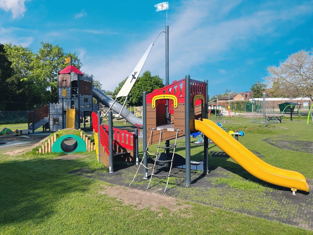 Priory Play Park features colourful play equipment, including a castle-themed climbing frame, slides, rope nets and swings. The playground is surrounded by open grassy areas and trees, with plenty of space for outdoor play.