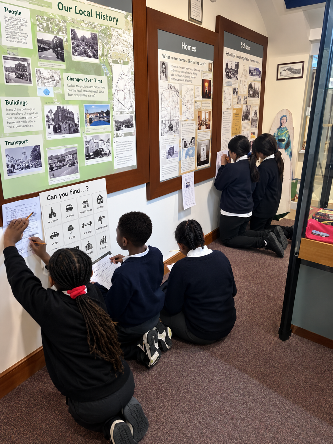 A group of schoolchildren in dark uniforms explore a museum exhibition, examining historical photographs, information boards and interactive displays. Some kneel on the carpet to look more closely at the exhibits.