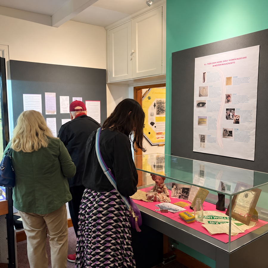 Three visitors explore a museum exhibition. One visitor examines historical objects and documents in a glass display case, while two others read information panels on the wall.