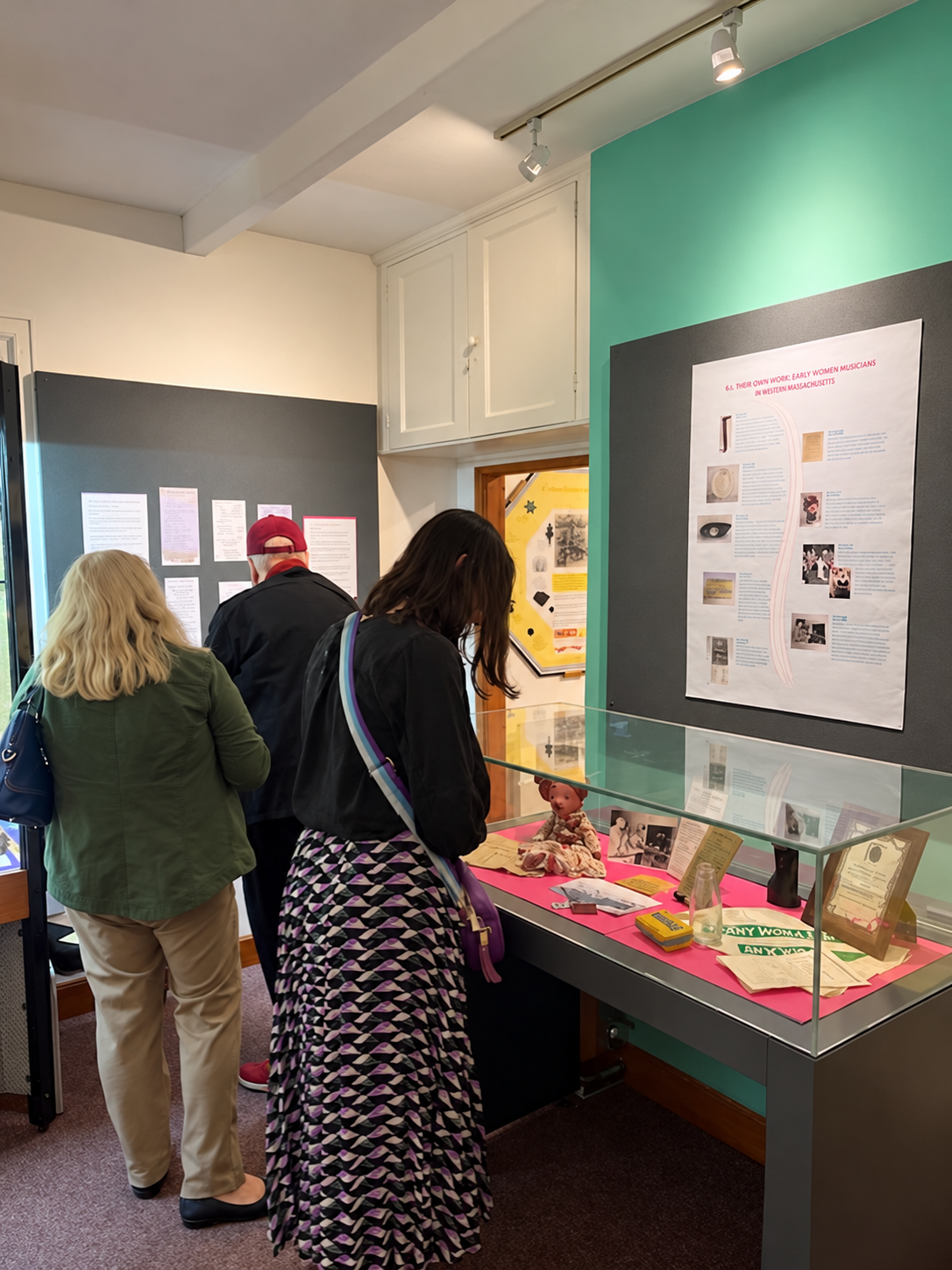 Three visitors explore a museum exhibition. One visitor examines historical objects and documents in a glass display case, while two others read information panels on the wall.