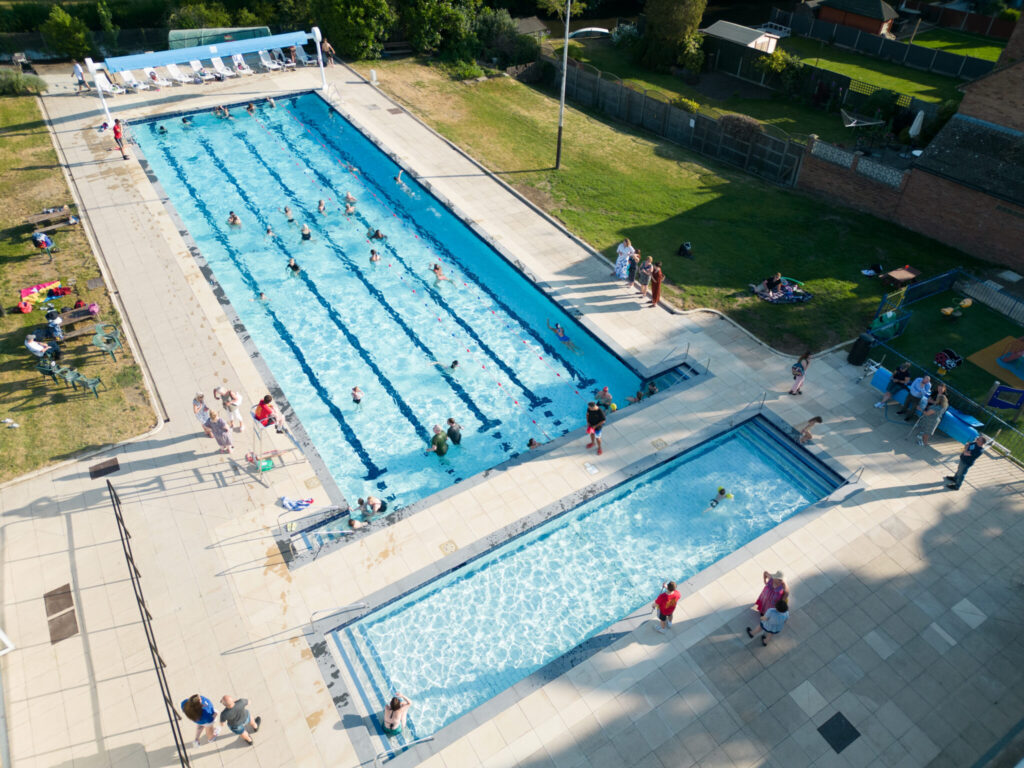 An aerial view of Ware Priory Lido on a sunny day, showing a large outdoor swimming pool and a smaller shallow pool, both with people swimming and enjoying the water. The pools are surrounded by paved areas, sun loungers and grassy spaces where visitors are relaxing.
